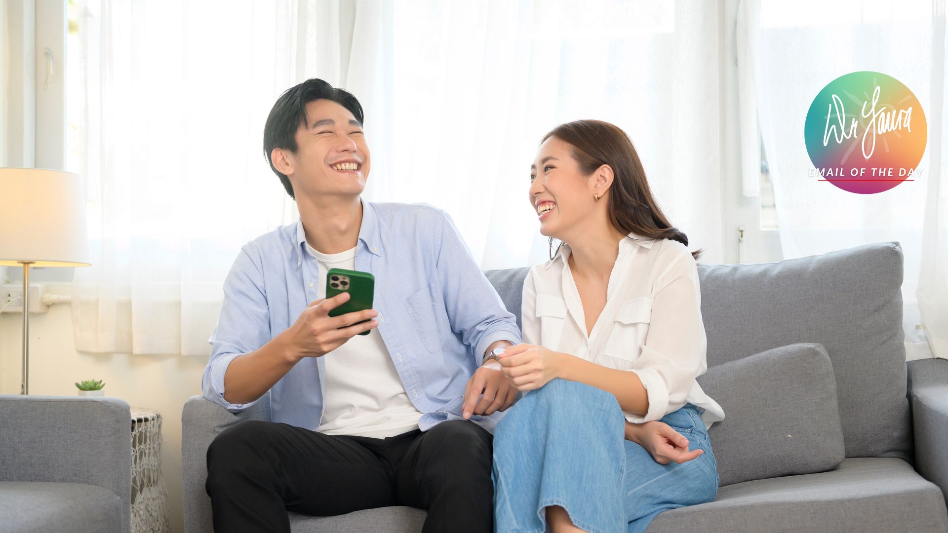 Man in blue button-up laughs while holds his phone and placing his arm on the legs of woman wearing white blouse