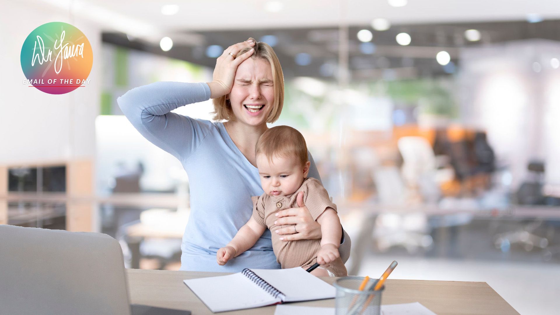Woman in blue shirt closes eyes and holds head in one hand while holding baby in beige onesie, sitting at a table with a notebook in front of her