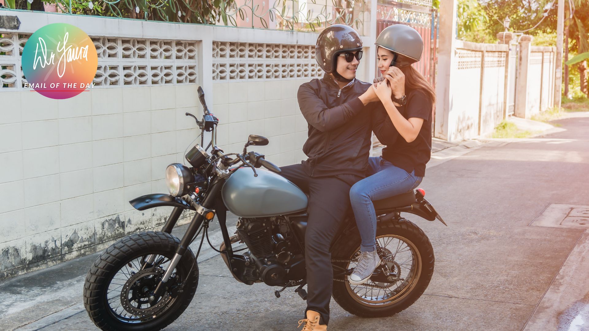 Man in black jacket and woman in black shirt sit next to each other on a parked motorcycle, holding hands