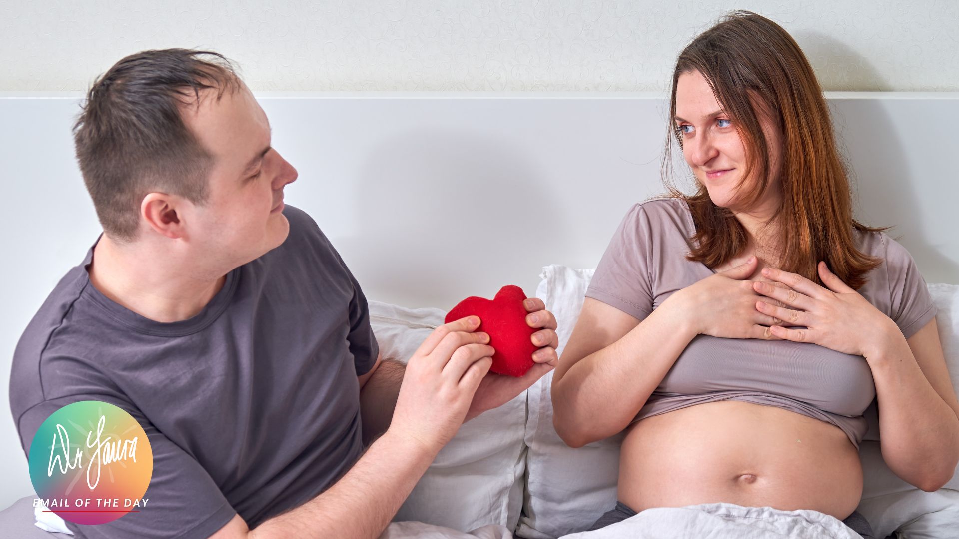 Woman in beige shirt with an exposed pregnant belly smiles at man sitting next to her in bed and offering a red heart plush