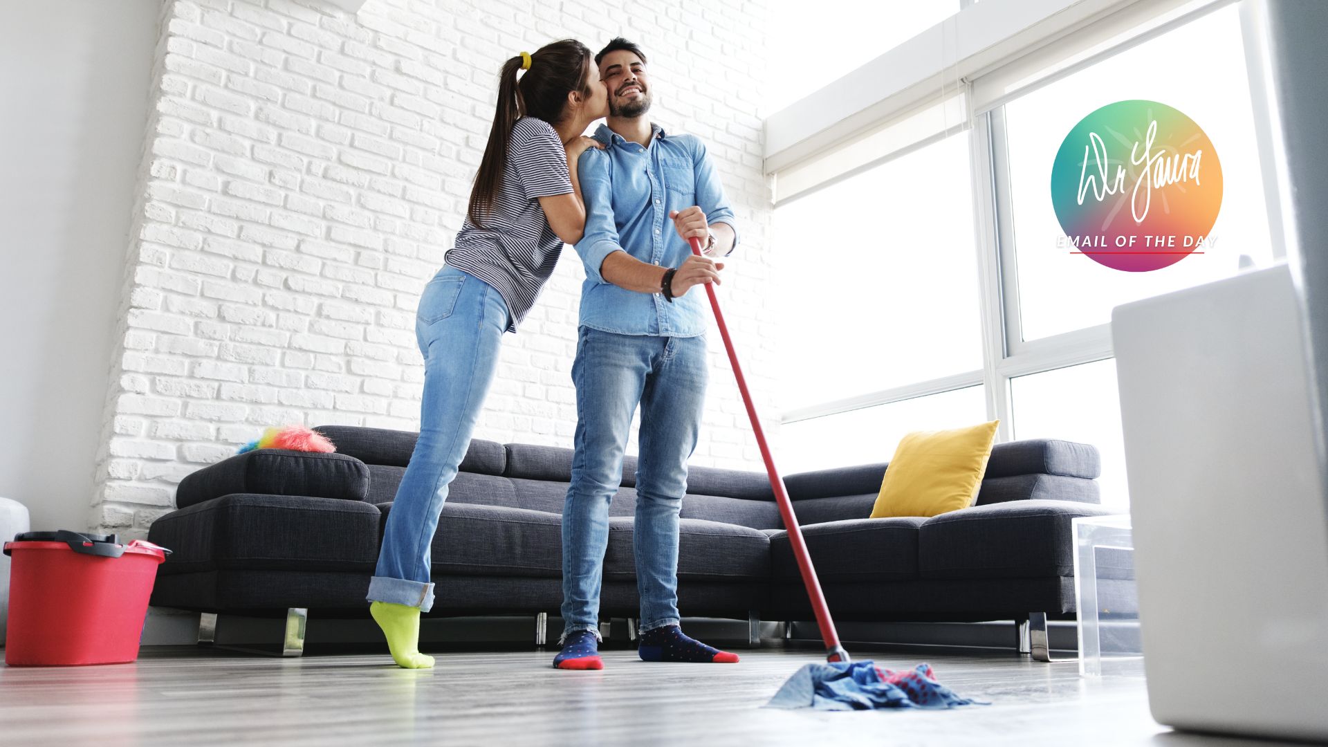 Woman in grey shirt stands on her toes to kiss man in blue button-up while he mops the floor