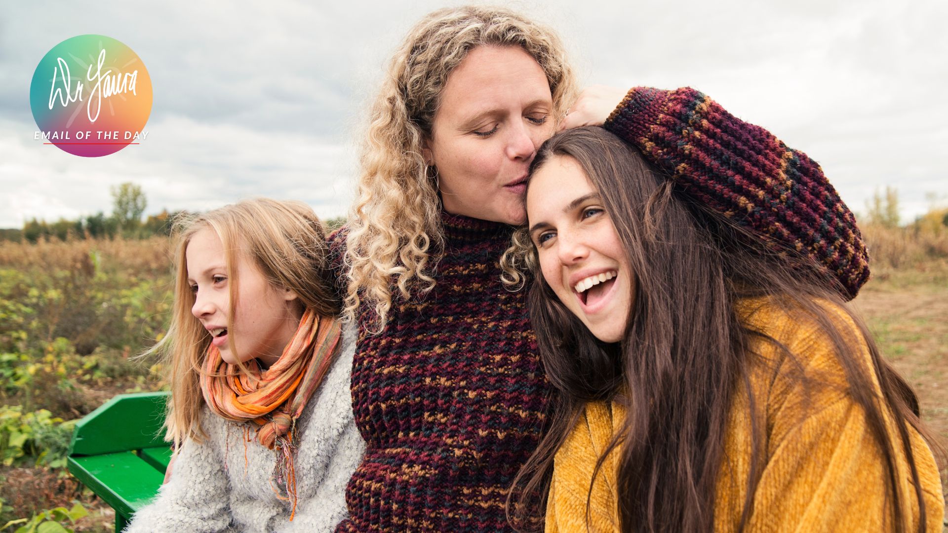 Woman in red sweater kisses the head of young female teen on her left and hugs a young girl on her right