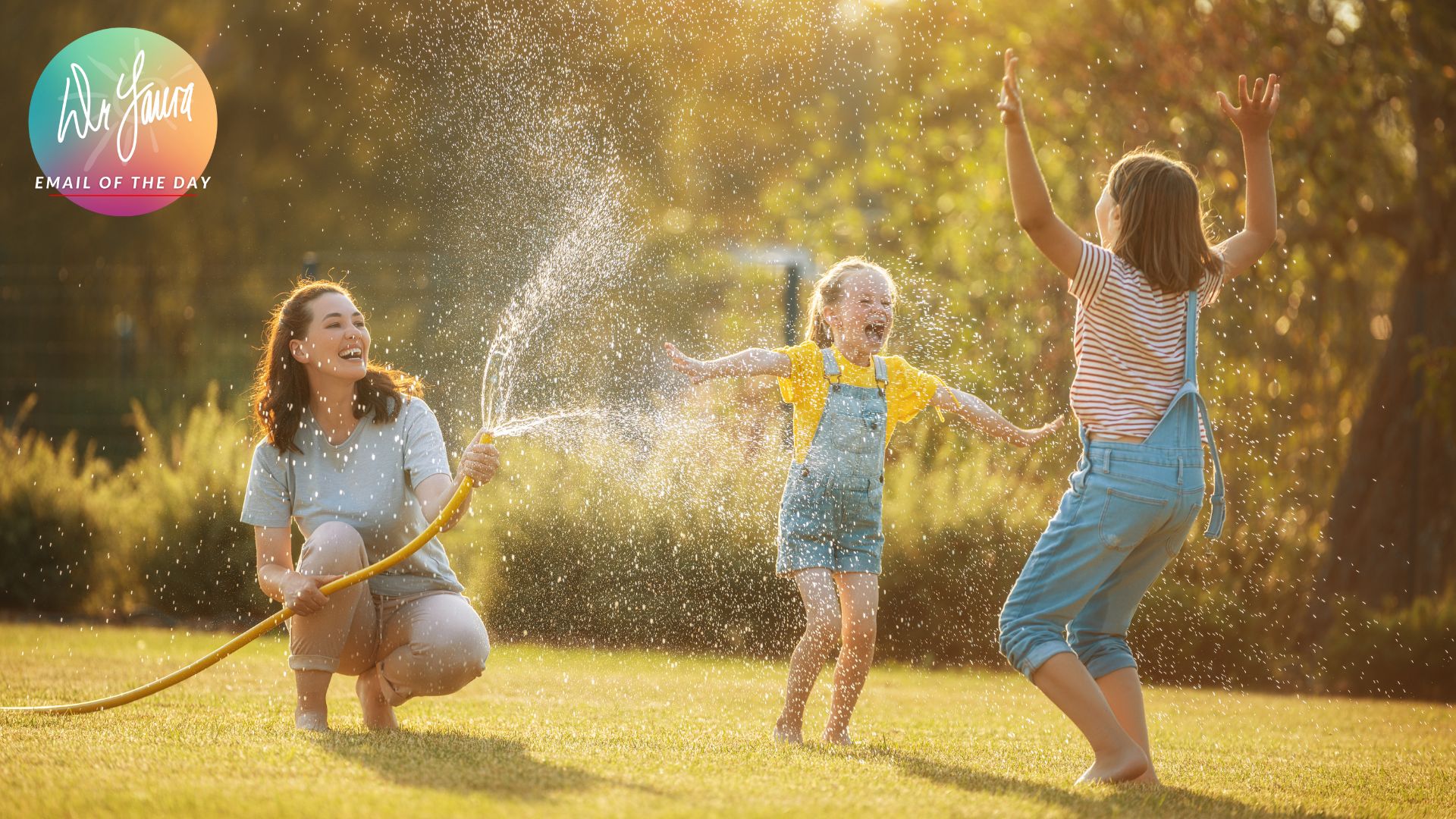 Woman in blue button-up sprays water from a hose on two girls in denim overalls