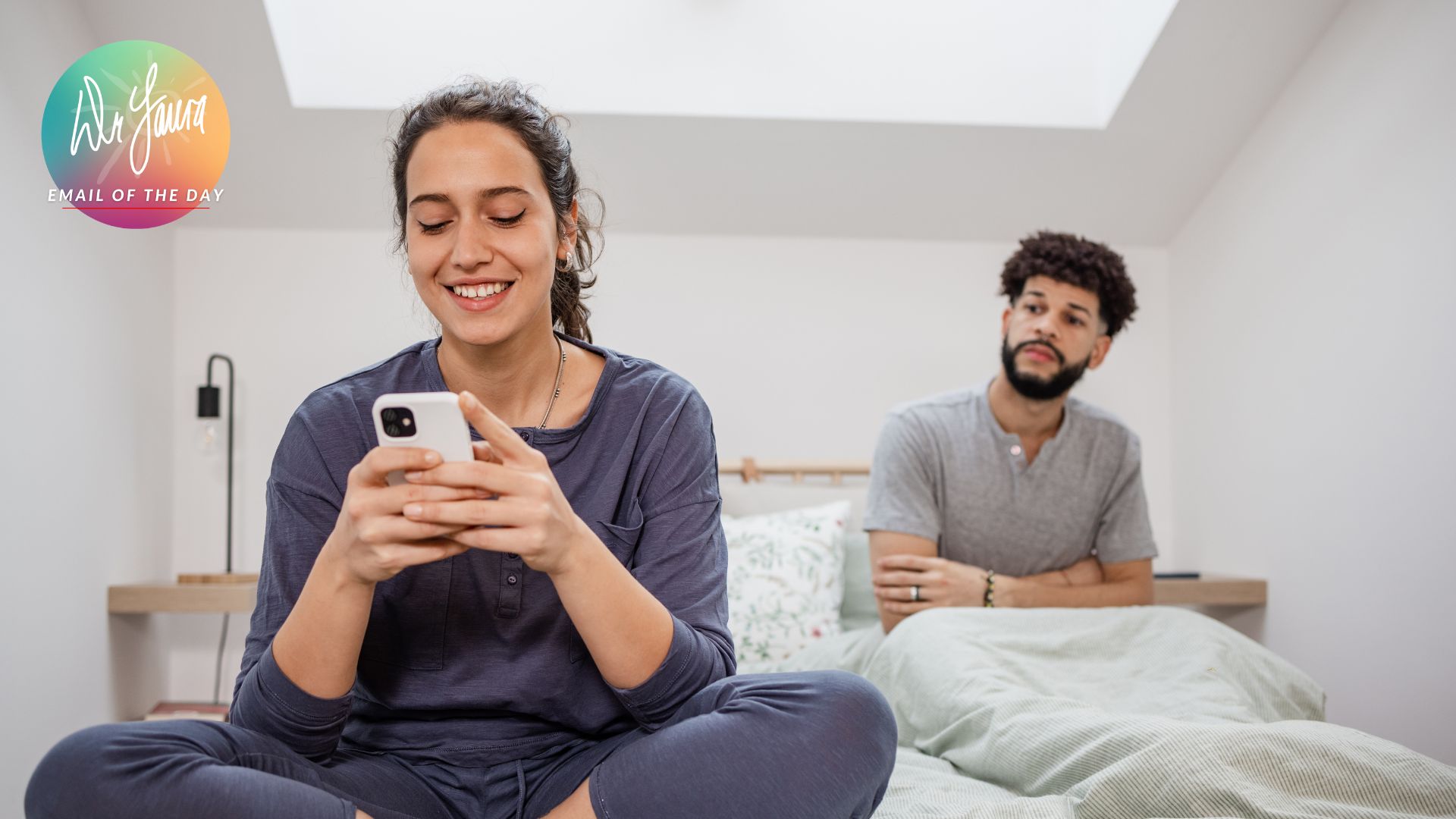 Woman smiles while looking at phone with man sitting behind her on the bed with his arms crossed and looking at her