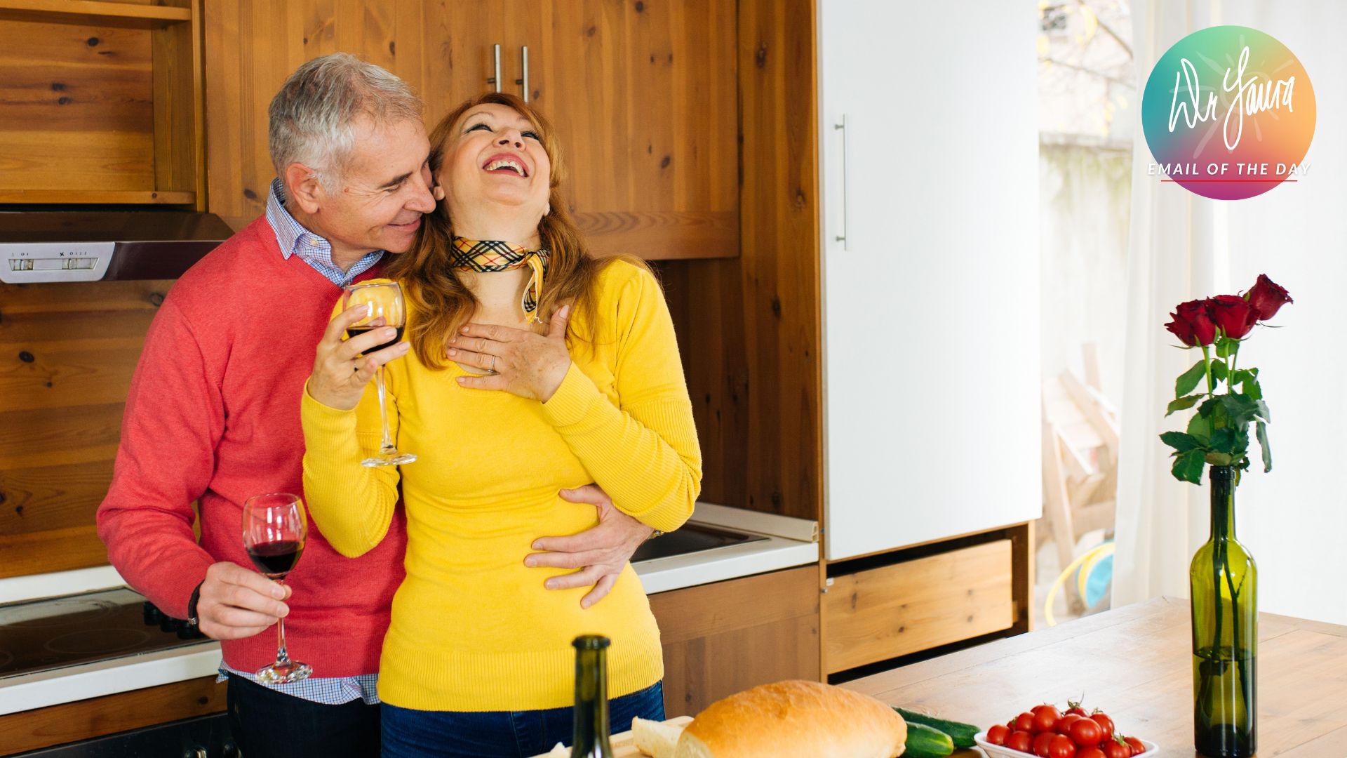 Man in red sweater embraces laughing woman from behind while they both hold wine glasses