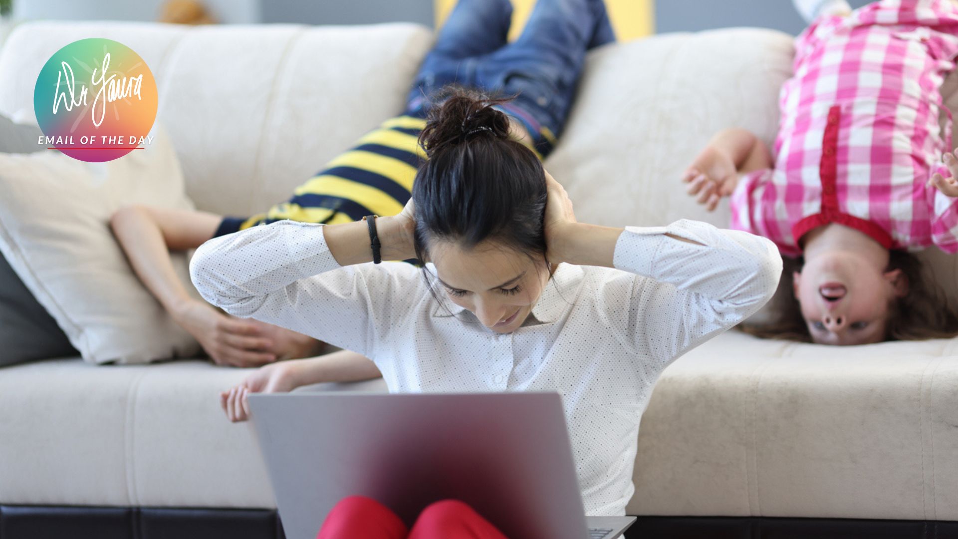 Woman covers her ears while sitting on the ground with a laptop on her lap, children sit on the couch behind her