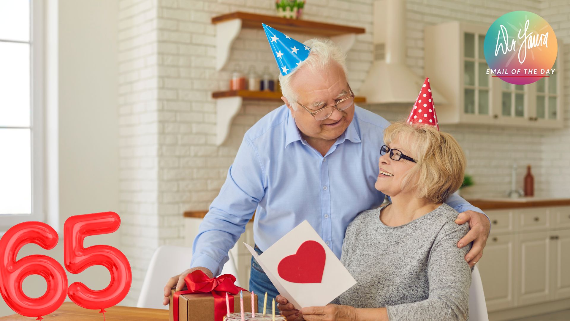 Man wearing birthday hat looks down at woman wearing birthday hat and opening card while sitting at table with a present, cake, and '65' balloons