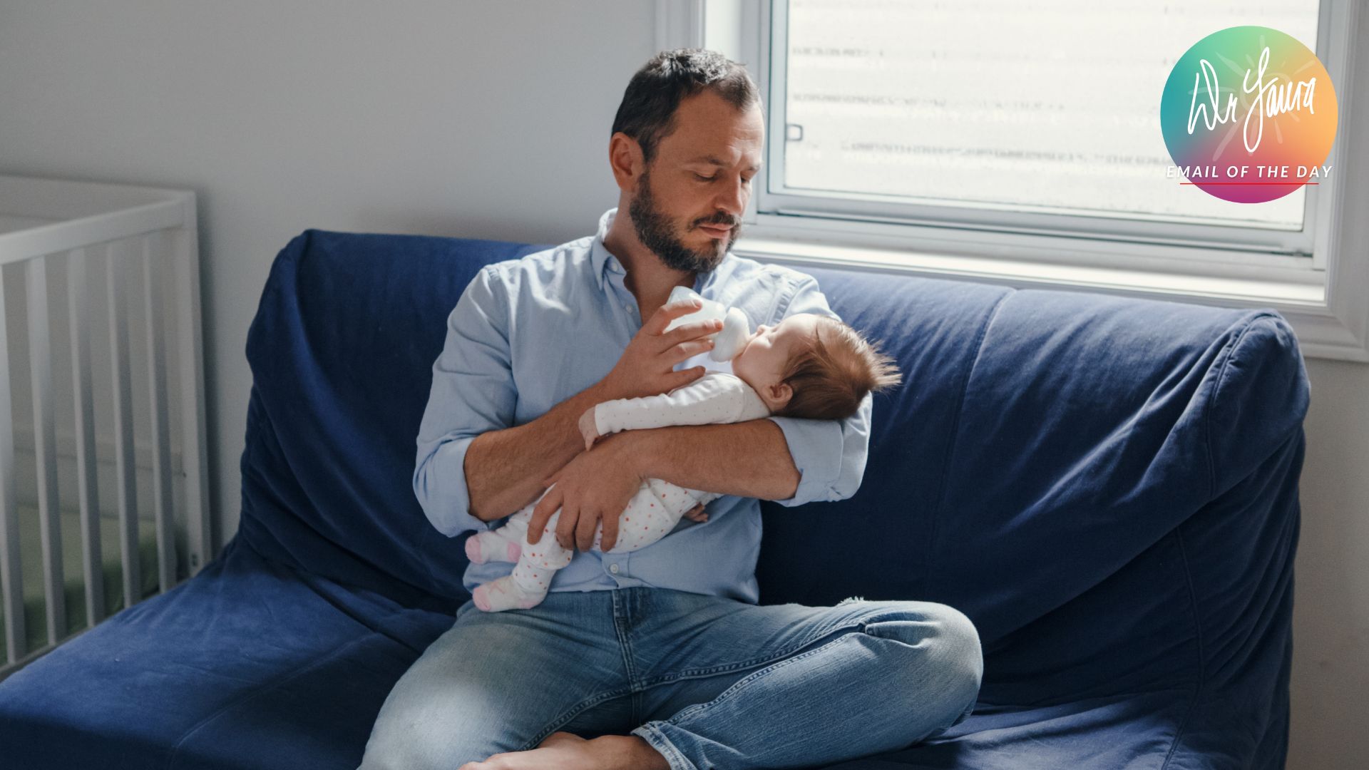 Man sits on couch and holds infant in his arm while feeding it a bottle