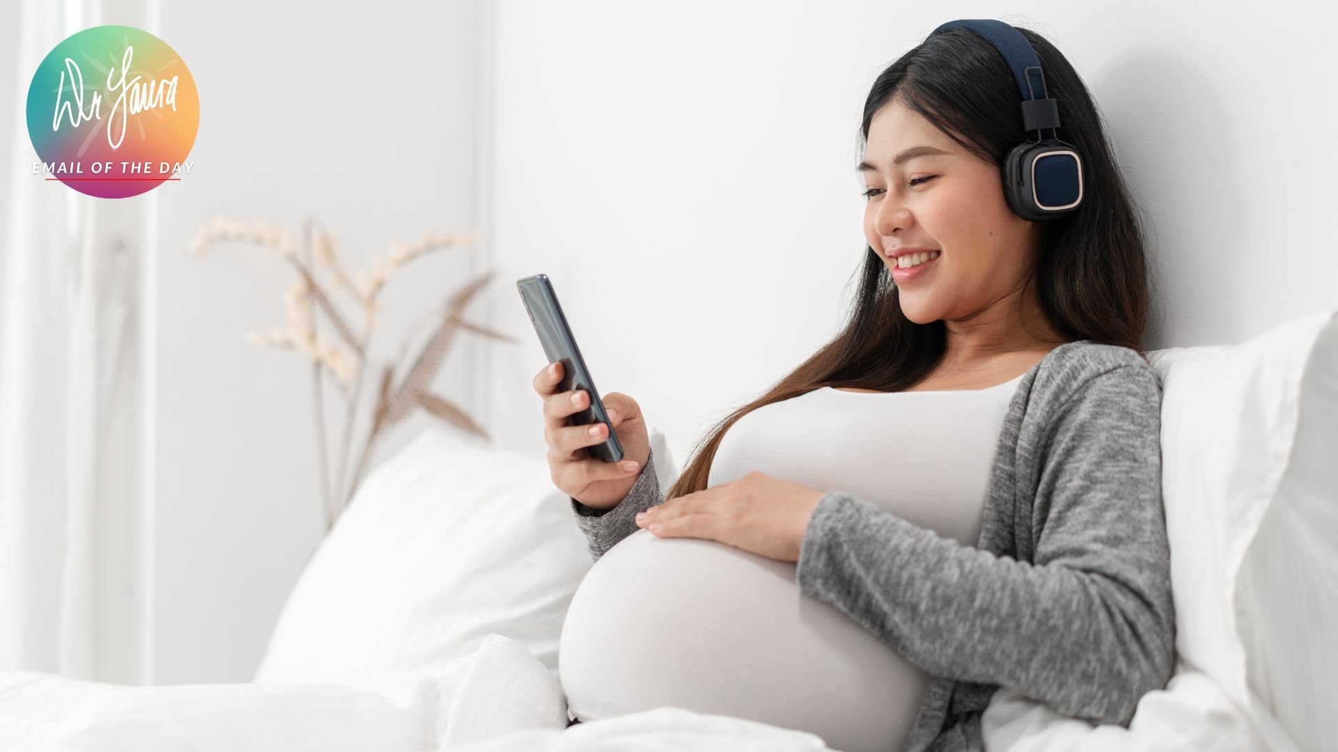 Woman wearing headphones smiles at phone while holding her baby belly