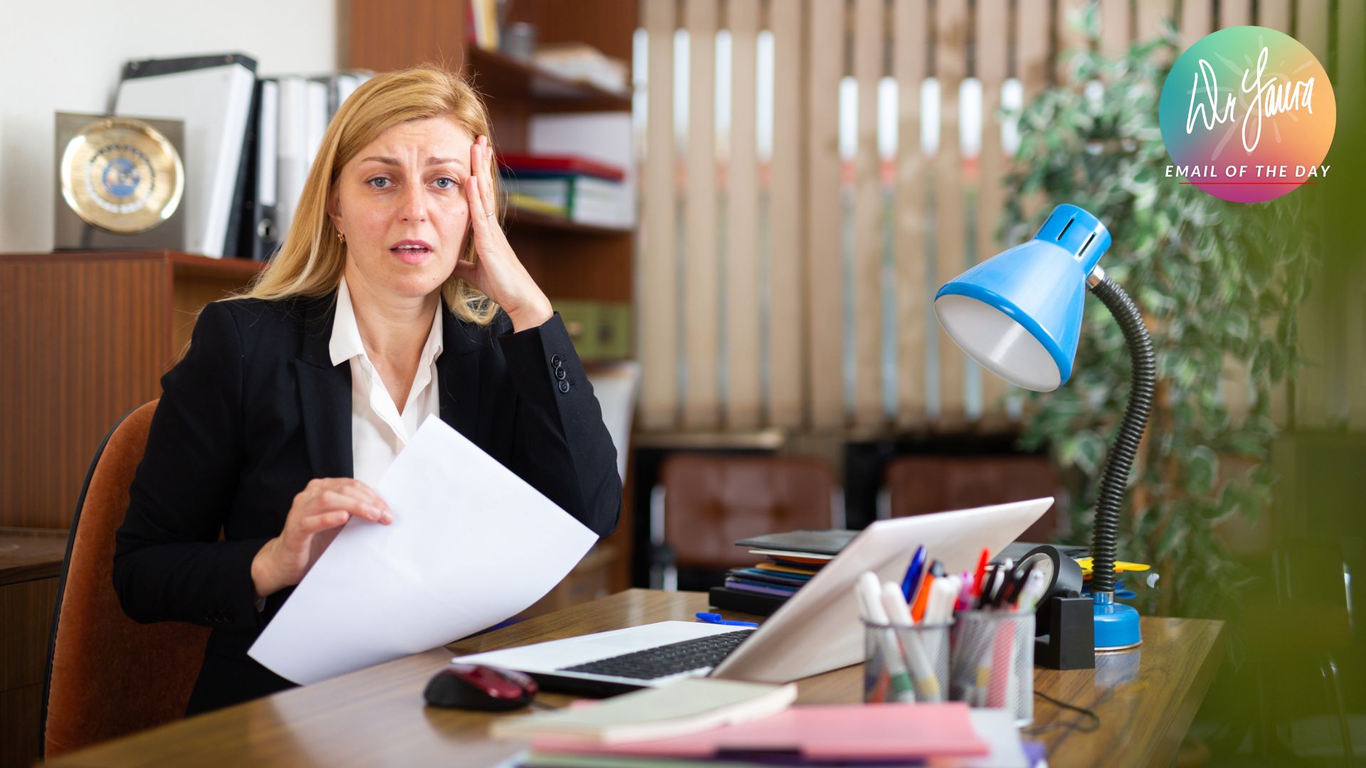 Woman looking stressed places hand on head while holding paper and sitting in front of work desk