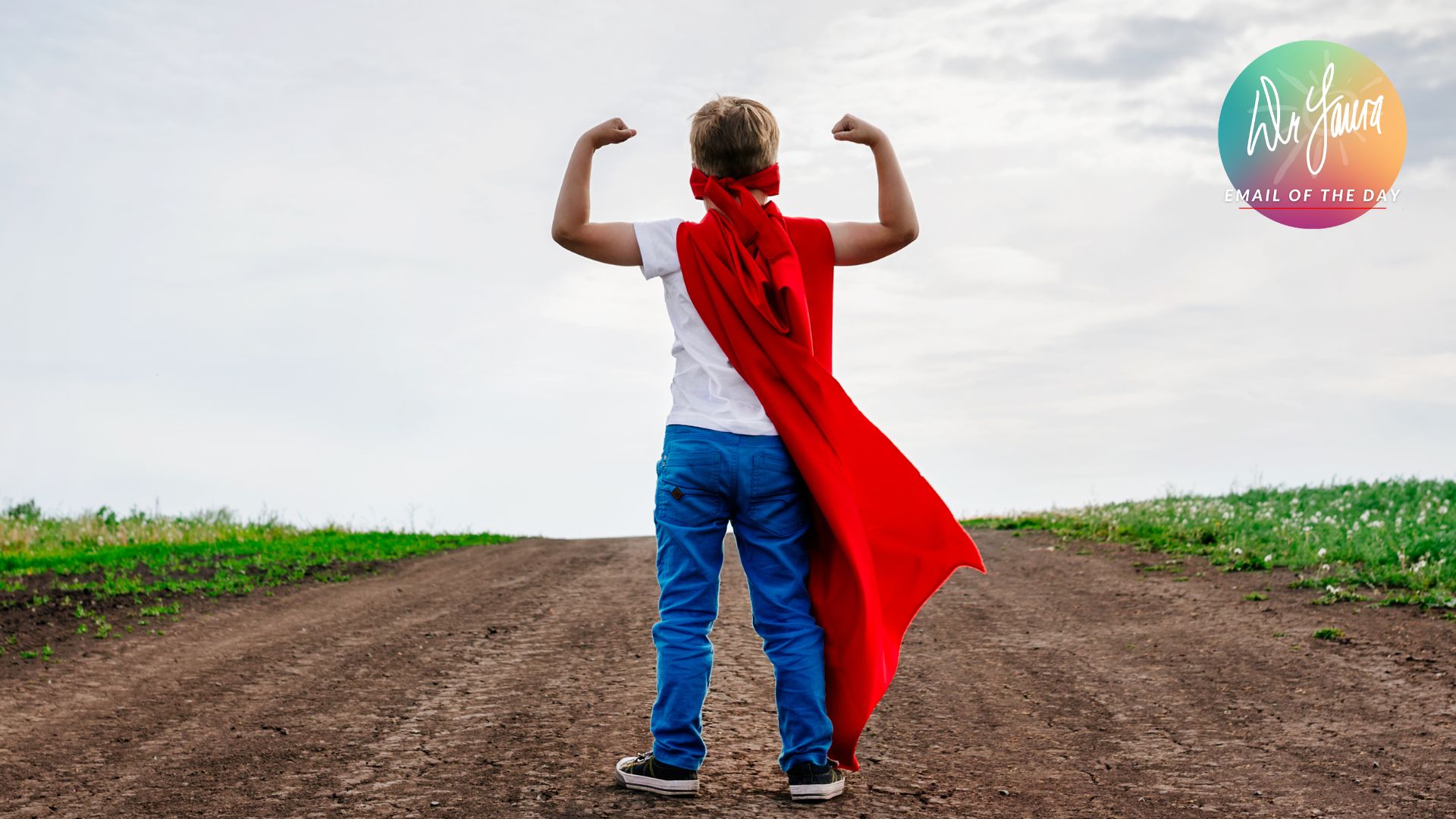 Boy in red cape flexes his biceps while standing in front of a dirt path in a field