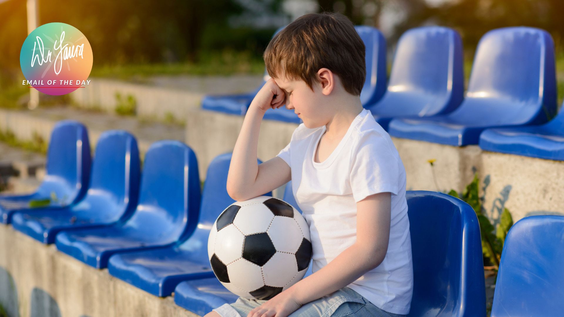 Little boy sits in stadium stands with a soccer ball on his lap and his fist resting on forehead