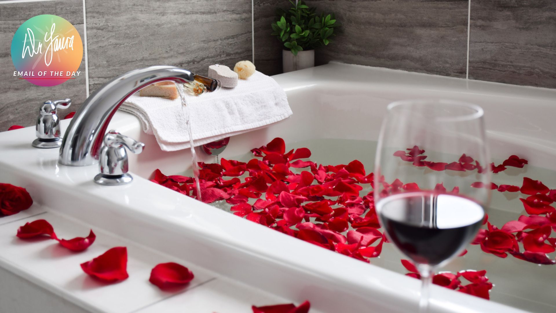 Bathtub is being filled with water while rose petals float on it and a wine glass with red wine sits next to bathtub