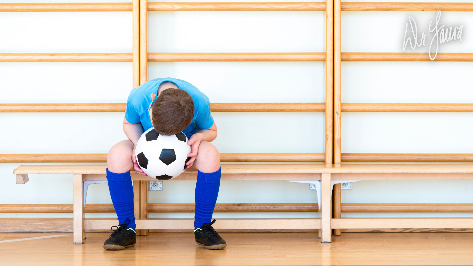 Boy in soccer uniform sits on bench while holding soccer ball and rests head on his knees